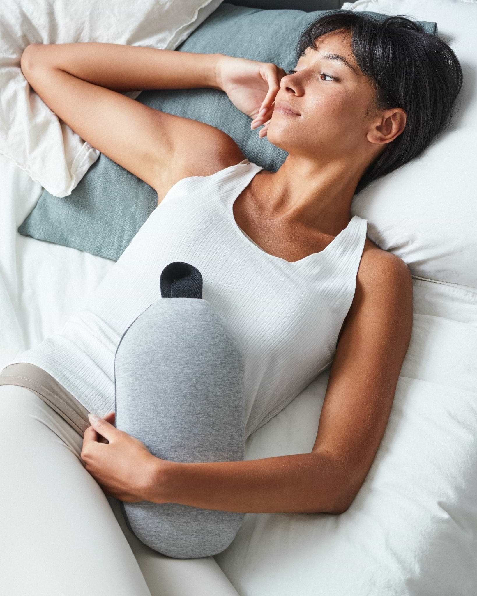 Close-up of a woman in a white t-shirt cradling a grey Ostrichpillow Heatbag under her arm against a clean grey background.