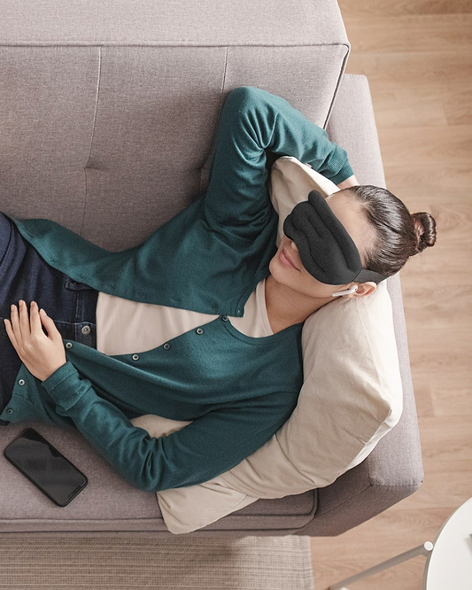 High-angle view of a woman lying comfortably on a grey fabric couch wearing a green cardigan and the cushioned Hot and Cold Eye Mask in Midnight Grey.