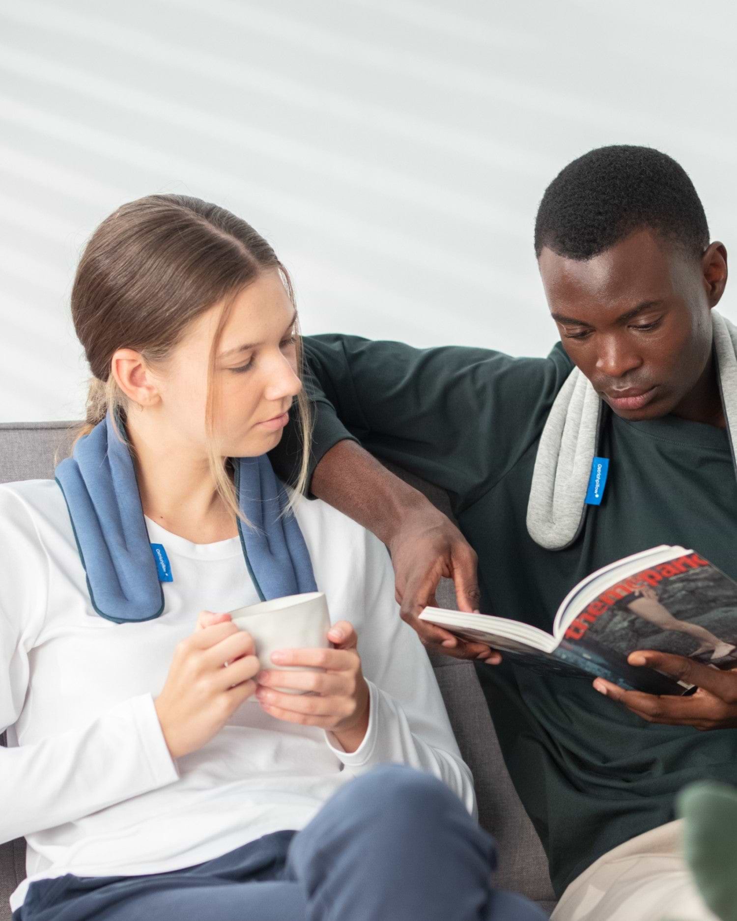 A woman wears the 'Ocean Blue' colorway for Heated Neck Wrap, sitting next to a man wearing the 'Midnight Grey' colorway.