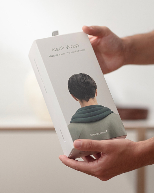 Close-up lifestyle and e-commerce packaging shot for the Heated Neck Wrap. A person holds the tall, rectangular white box.