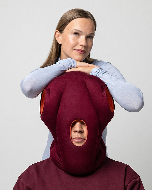 A woman rests her arms and chin on top of the 'Spiced Plum' pillow, which completely covers another person's head, framing only their nose and mouth.