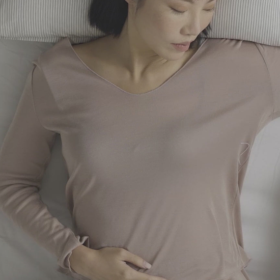 Woman lying down in bed using the Ostrichpillow Heatbag clay heating pad for stomach cramps and relaxation.