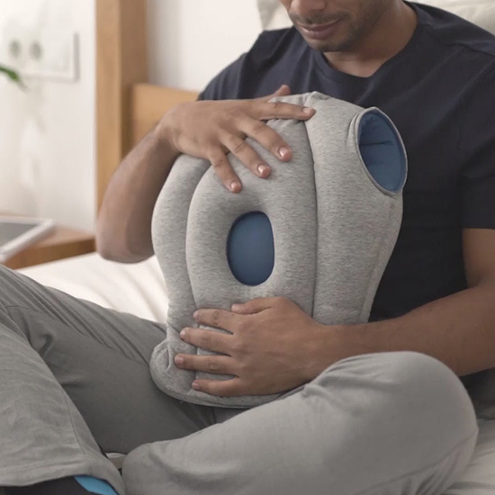A man demonstrates how to wear Original Napping Pillow. He pulls it over his head and ensures his nose and mouth align with the breathing opening.