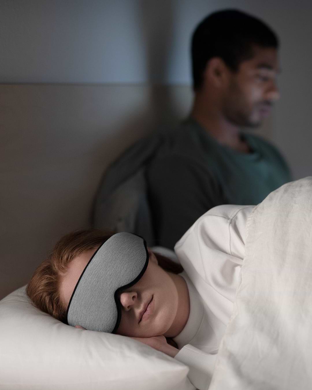 A woman sleeps wearing a Midnight Grey Eye Mask that blocks out light and a man shows how the mask enables undisturbed sleep.