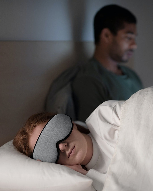 A woman sleeps wearing a Midnight Grey Eye Mask that blocks out light and a man shows how the mask enables undisturbed sleep.