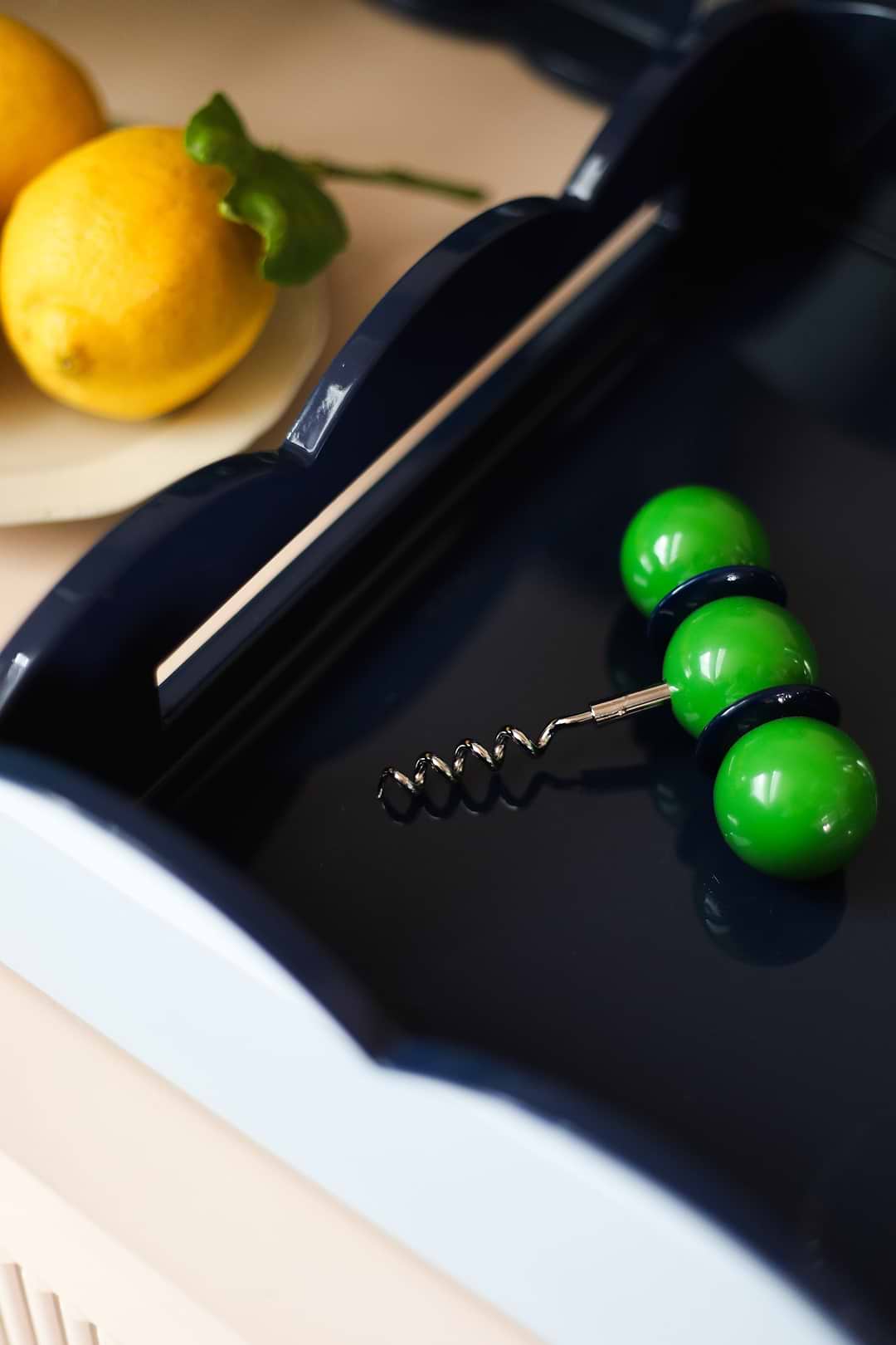 Close-up of an Addison Ross green ball corkscrew resting inside a navy scalloped lacquer tray beside fresh lemons