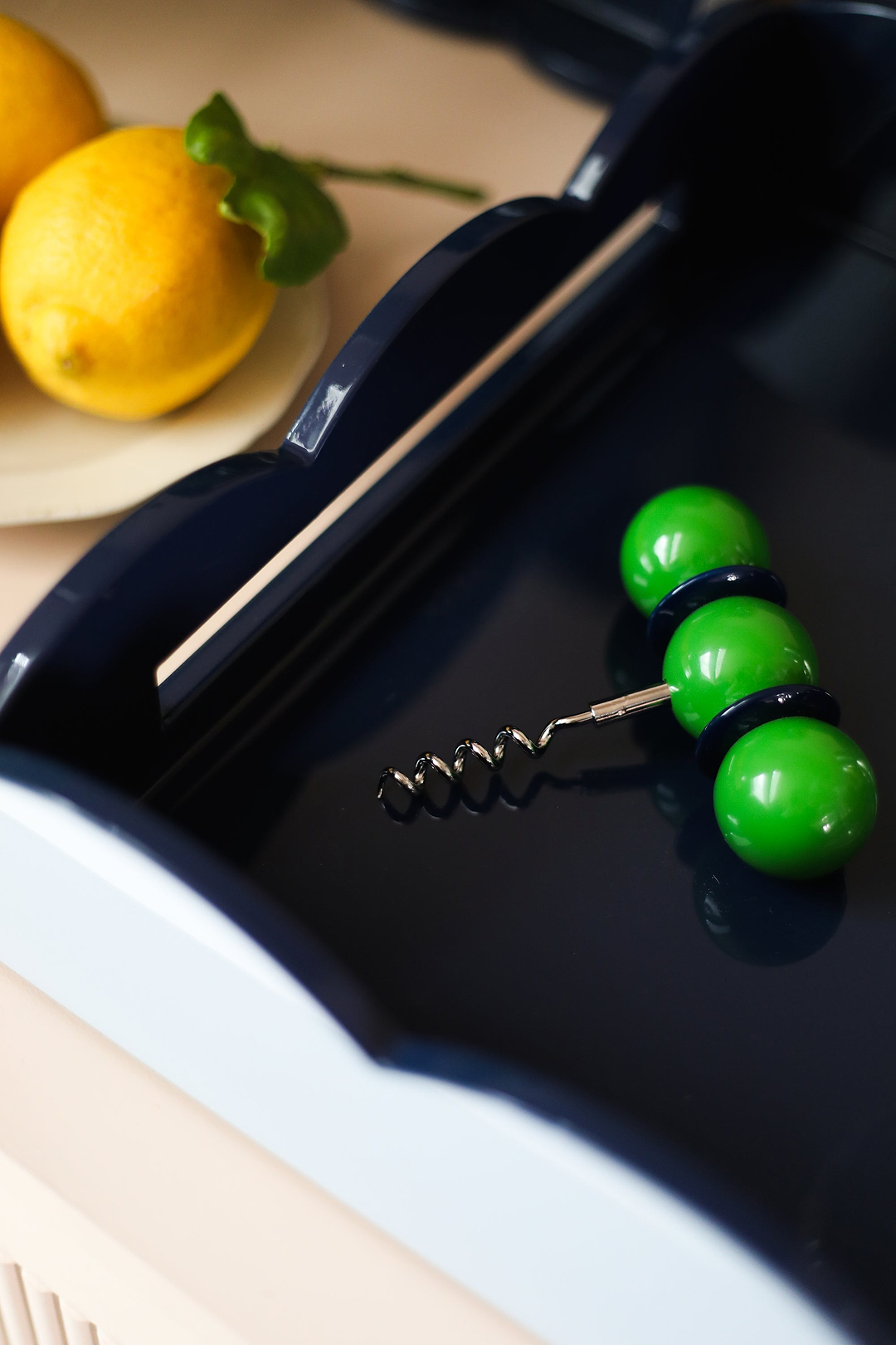 Close-up of an Addison Ross green ball corkscrew resting inside a navy scalloped lacquer tray beside fresh lemons