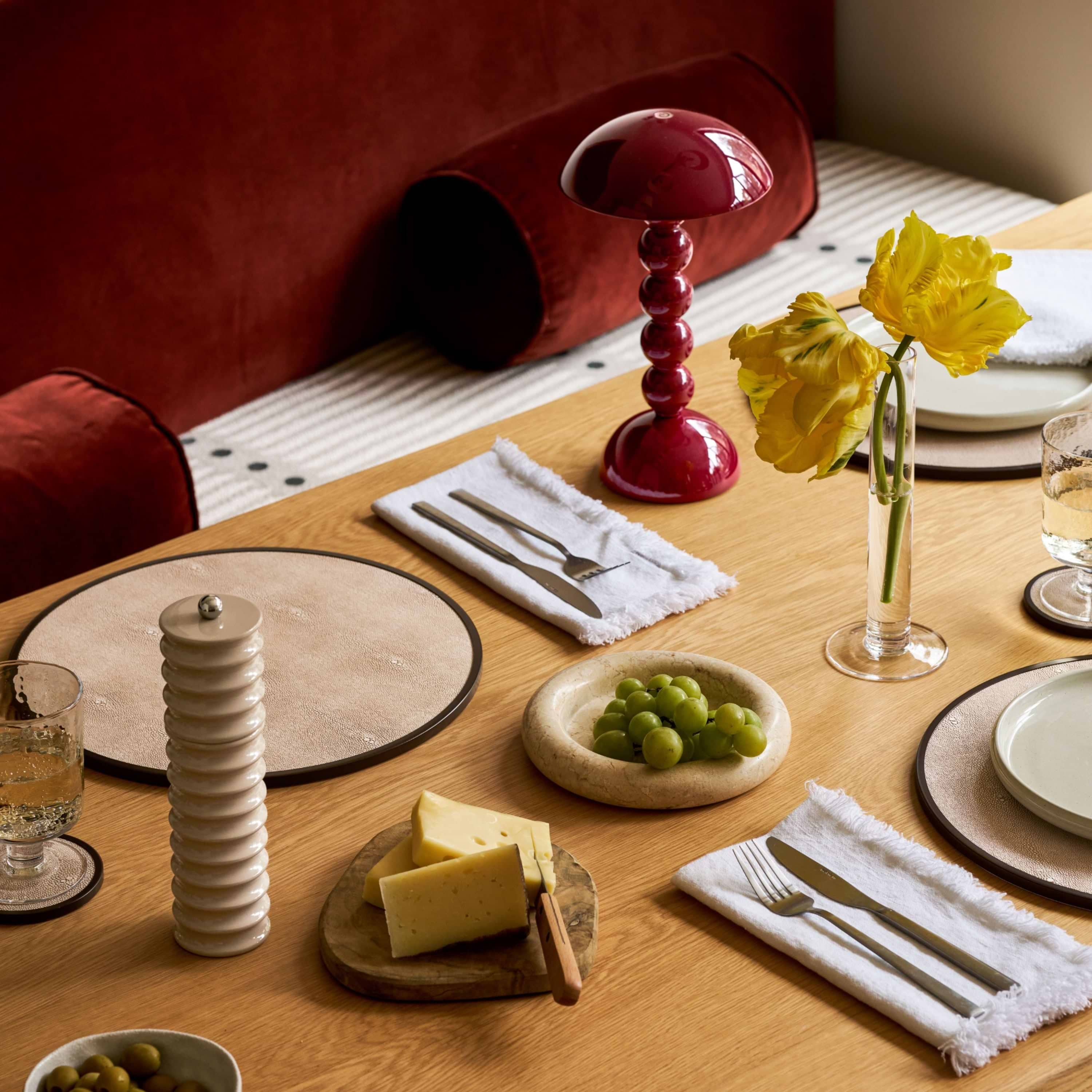 A styled dining table setting with white plates, white napkins, silver cutlery, a small bowl of green grapes, cheese on a wooden board, a white ceramic pepper mill, and a clear glass vase holding yellow flowers. A red mushroom-shaped table lamp and deep red upholstered seating are visible in the background.