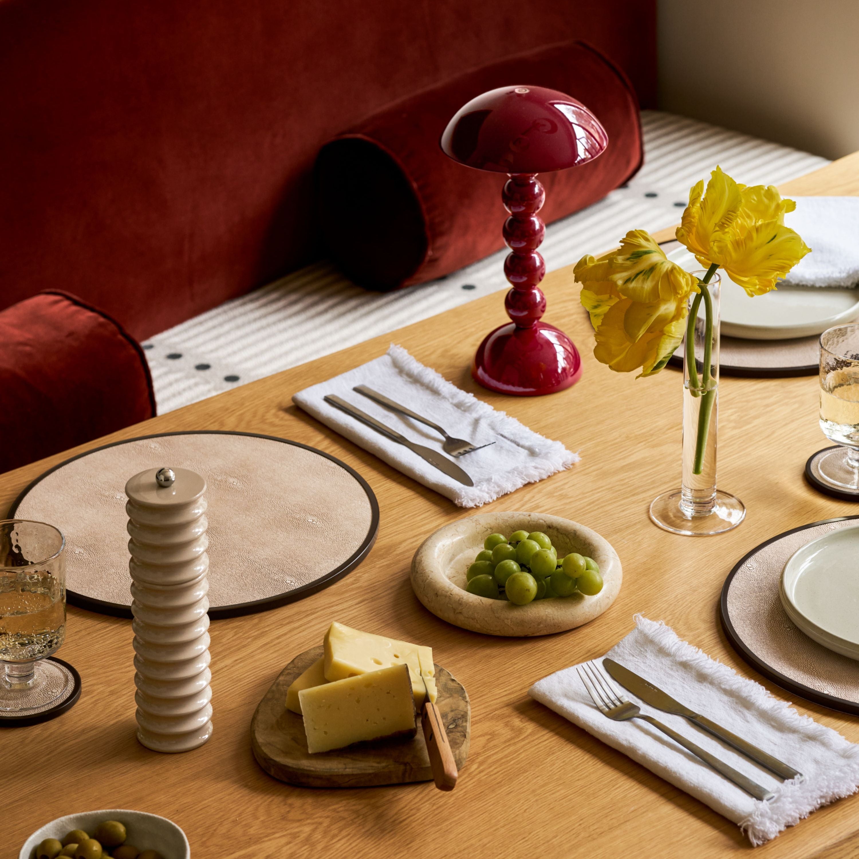 A styled dining table setting with white plates, white napkins, silver cutlery, a small bowl of green grapes, cheese on a wooden board, a white ceramic pepper mill, and a clear glass vase holding yellow flowers. A red mushroom-shaped table lamp and deep red upholstered seating are visible in the background.