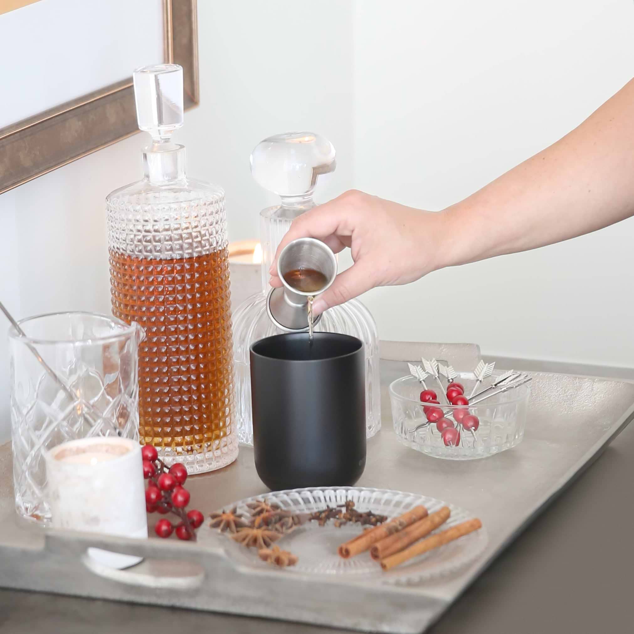 A hand pours liquor into a black Ember Mug² that's surrounded with ornate glassware, cinnamon sticks, cloves, star anise, and cranberries.