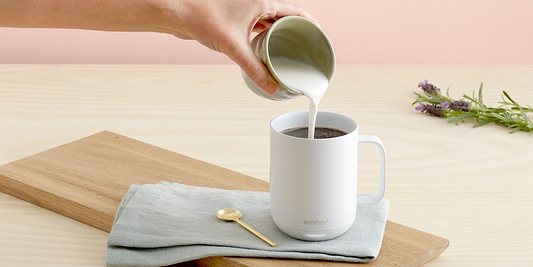 A hand pours cream from a small cup into a white Ember Mug² that's sitting on a blue cloth which is neatly folded and resting on a wooden cutting board. Next to the mug is a gold spoon, and there is a branch of green eucalyptus leaves in the background.