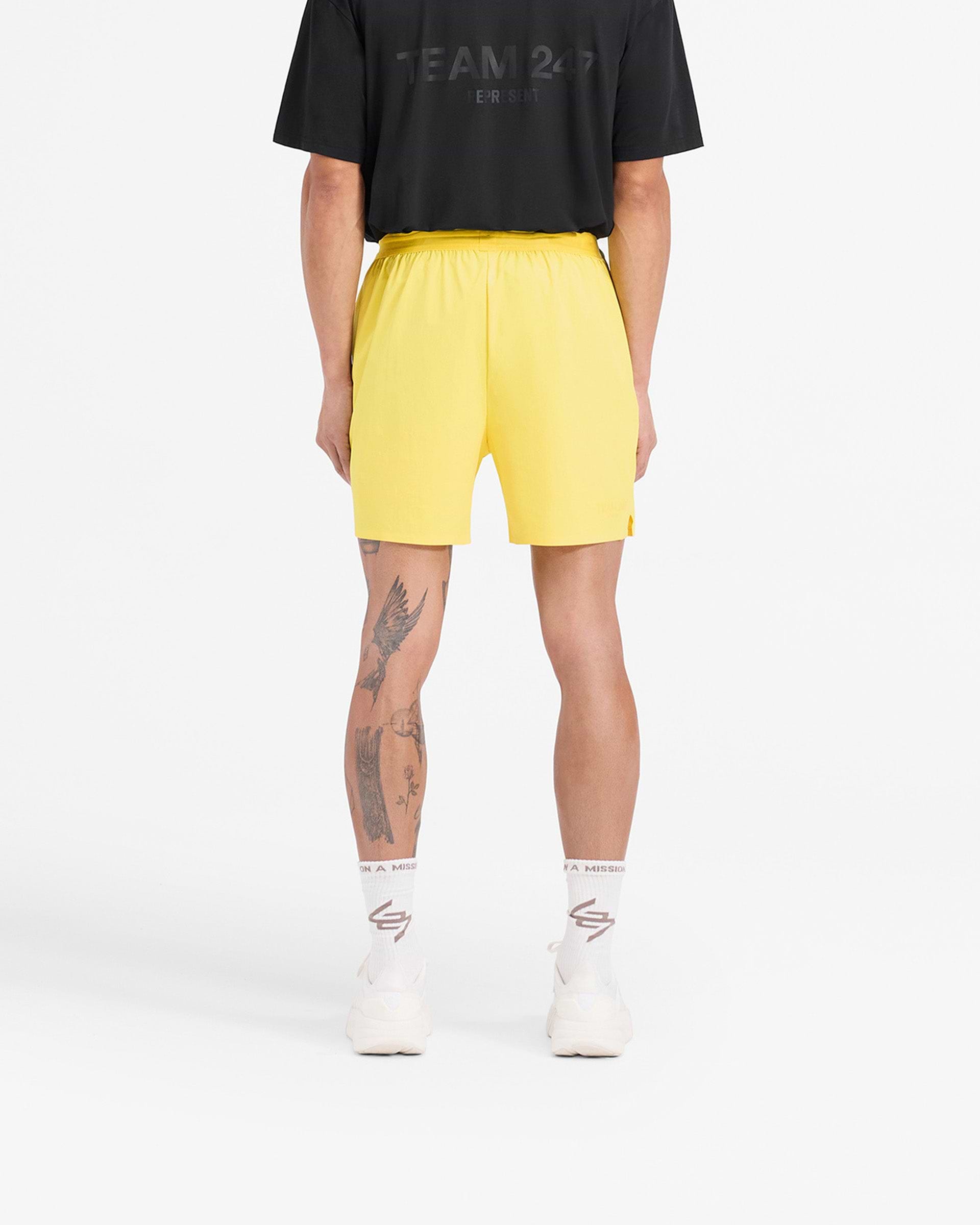 Man wearing yellow shorts on a plain white background | Team 247 Fused Shorts in Lemon | Gym shorts with quick drying fabric and 247 branding | 247 Shorts | Represent Clo