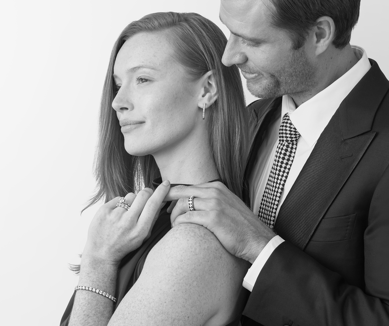 A black and white lifestyle photograph of a man in a suit and tie looking down at a woman who is looking to the side. She is wearing a lab-grown diamond necklace, bracelet, and ring.