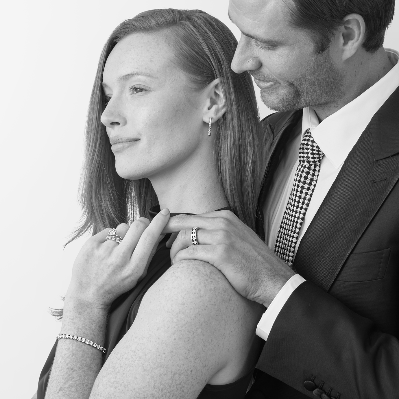 A black and white lifestyle photograph of a man in a suit and tie holding a woman from behind, gently touching her shoulder and neck where she is wearing lan grown diamond jewelry, including a lab-grown diamond ring, bracelet, and earring.