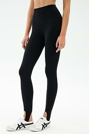 A person wears the SPLITS59 River High Waist Airweight Stirrup legging in black with white athletic shoes, standing against a plain light background.