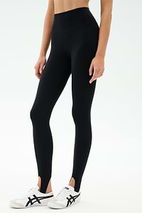 A person wears the SPLITS59 River High Waist Airweight Stirrup legging in black with white athletic shoes, standing against a plain light background.