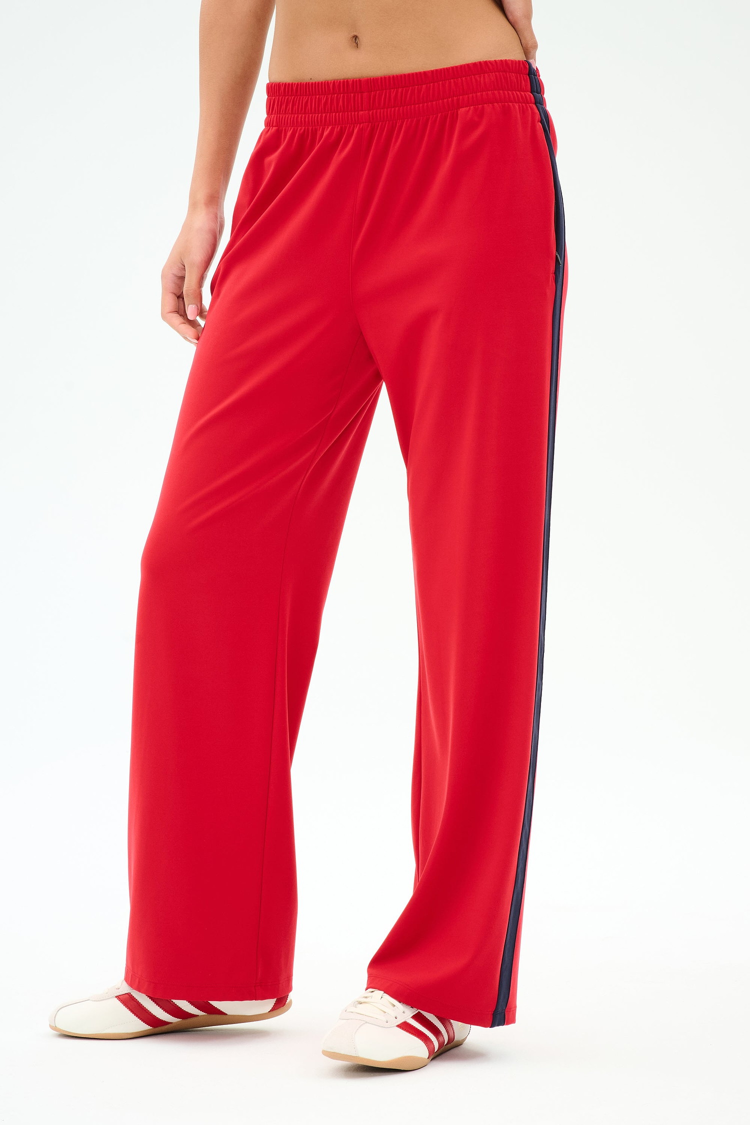 A person wearing SPLITS59 Maxie Rigor Track Pant in Pirate Red/Indigo, paired with white sneakers featuring red accents, stands against a plain background.