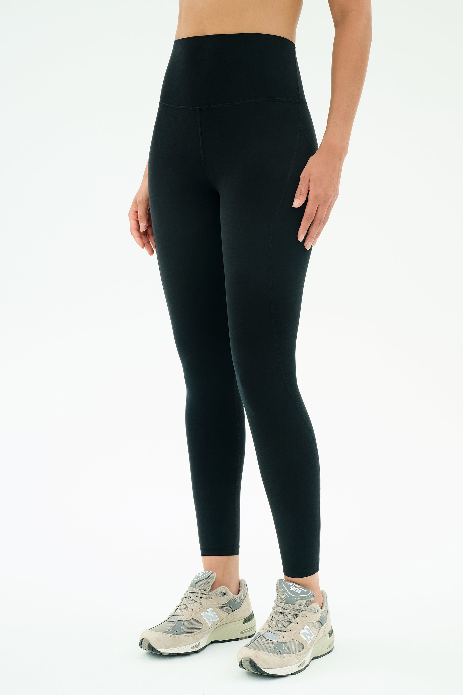 A person wears SPLITS59 Airweight High Waist 7/8 Pocket Legging in black and gray New Balance sneakers, standing against a plain white background.
