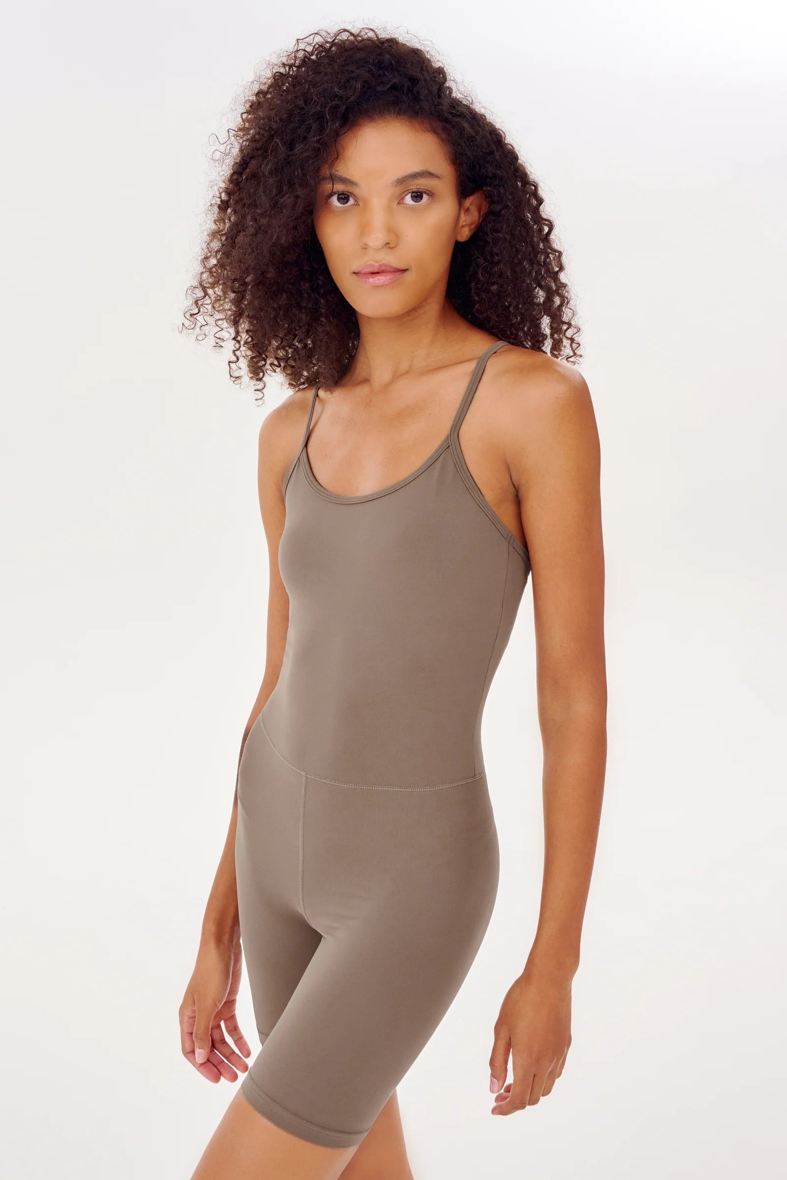 A person stands against a plain background, wearing a taupe-colored fitted sleeveless romper with thin straps and bike shorts-length legs.