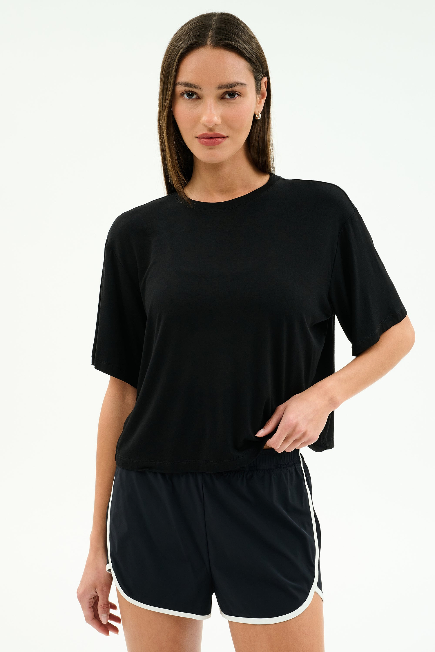 A woman stands against a plain background wearing the SPLITS59 Djuna Oversized Jersey Tee in black and black athletic shorts with white trim, perfect for gym workouts.