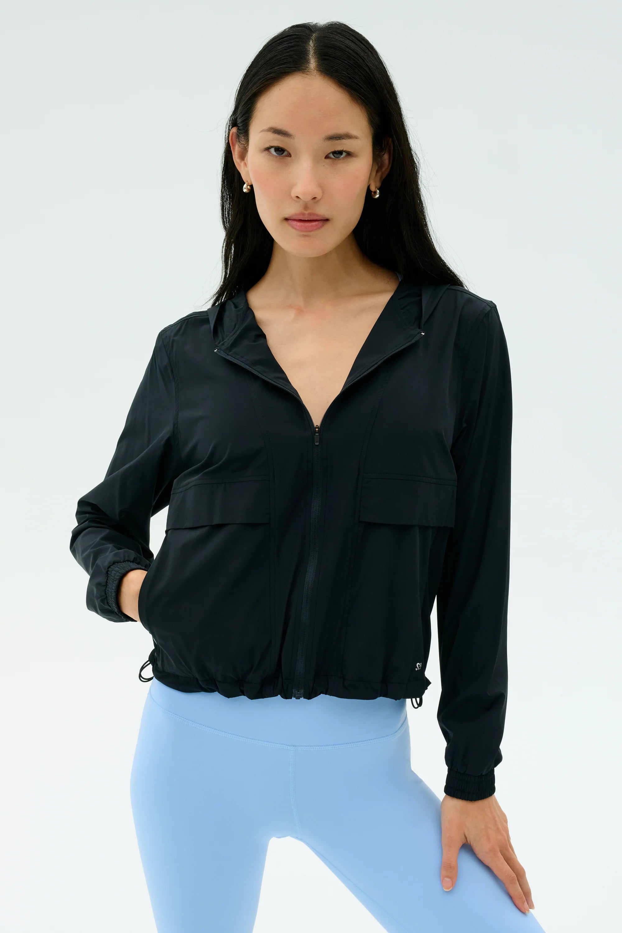 A woman wearing a black zip-up jacket with front pockets and light blue leggings stands against a plain light background.