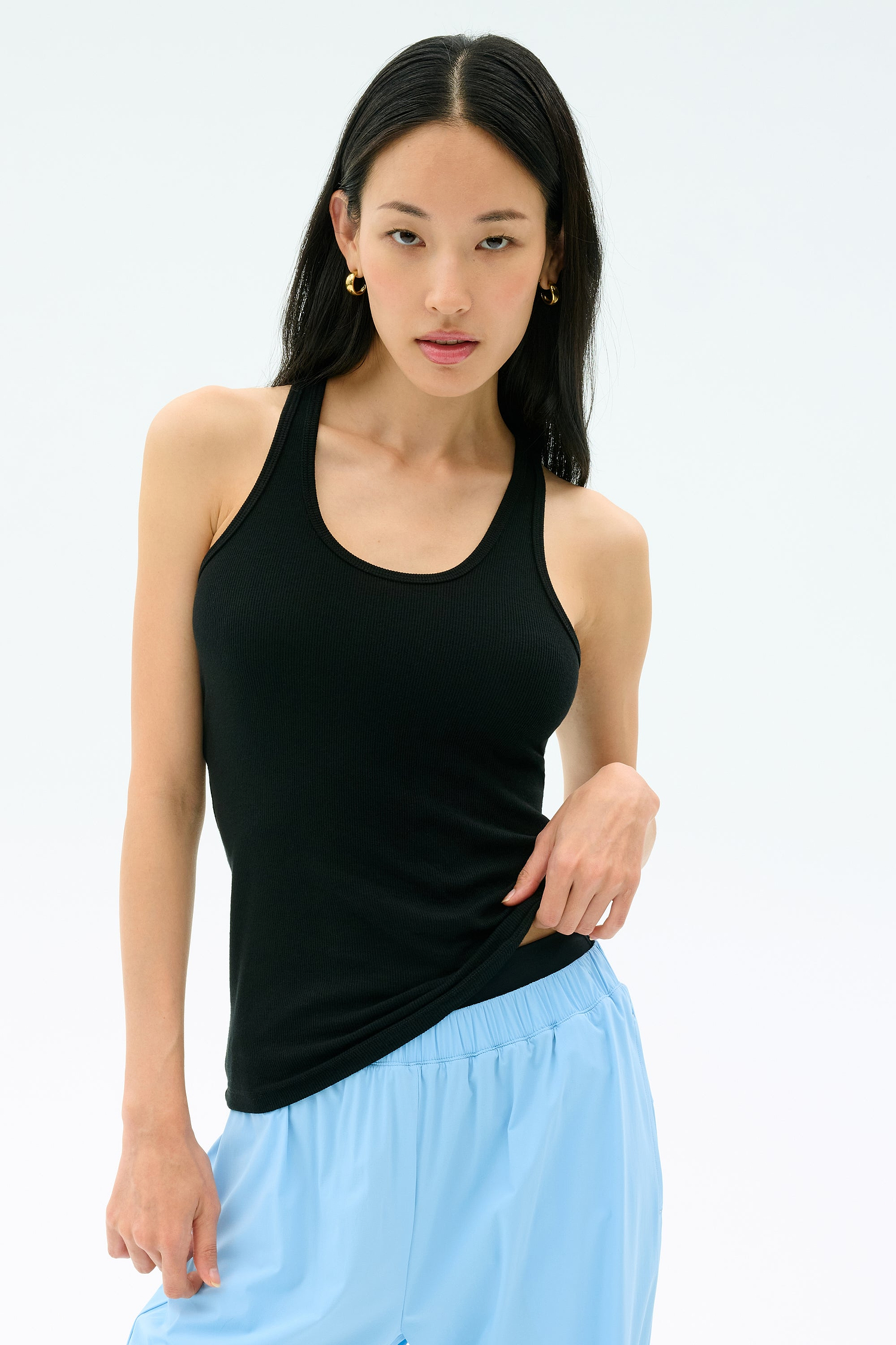 A woman with long black hair wears the Splits59 Ashby Rib Tank in black with light blue pants, standing against a plain light background.