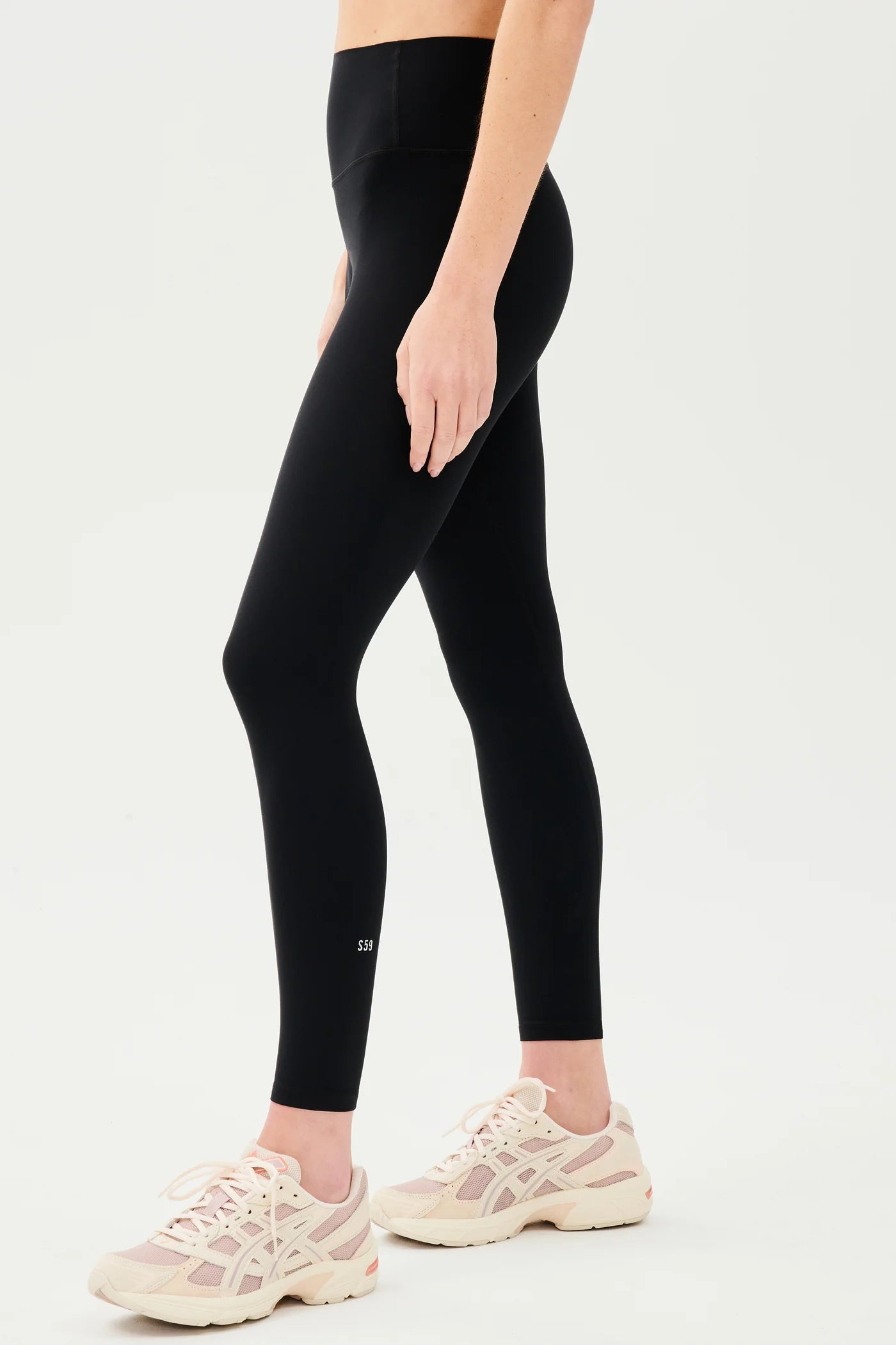 Person wearing high-waisted black leggings and light pink sneakers, standing sideways against a plain white background.