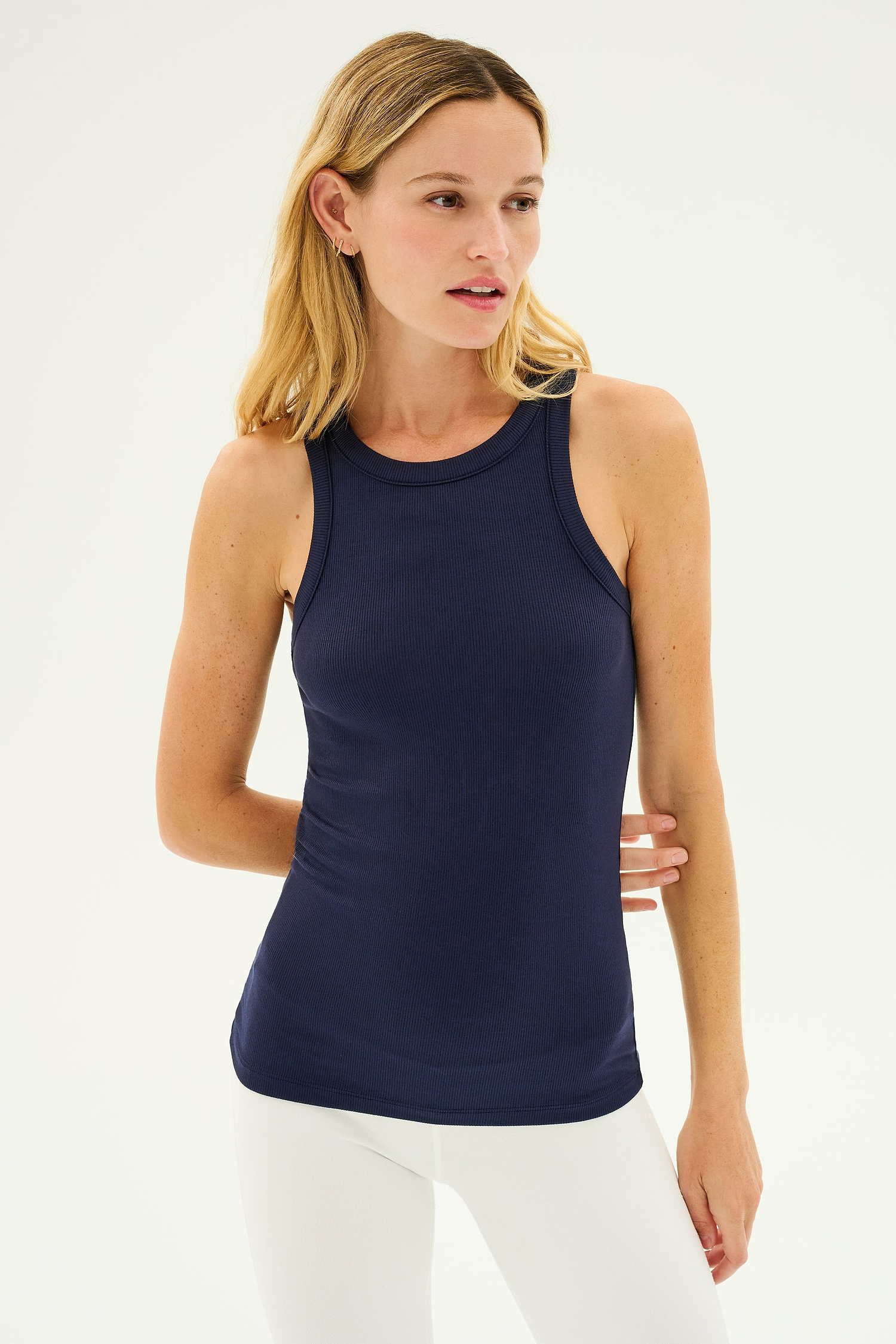 A woman with long blonde hair wears the SPLITS59 Kiki Rib Tank Full Length in Indigo, featuring a high racer neckline, paired with white leggings. She is looking to her right with a neutral expression against a plain background, evoking the essence of dedicated gym workouts.