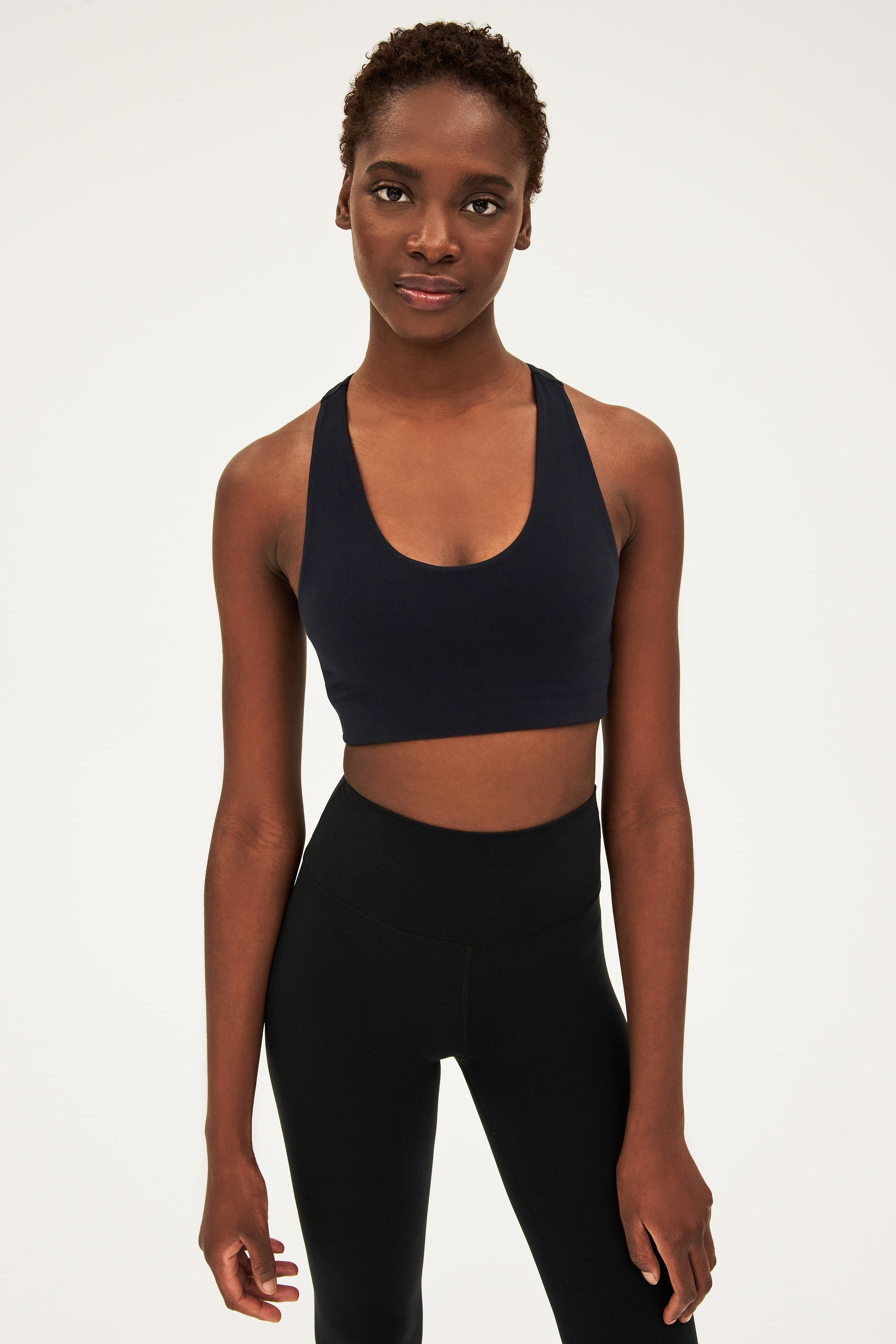 A person stands facing the camera, wearing a black sports bra and black high-waisted leggings against a plain, light background.