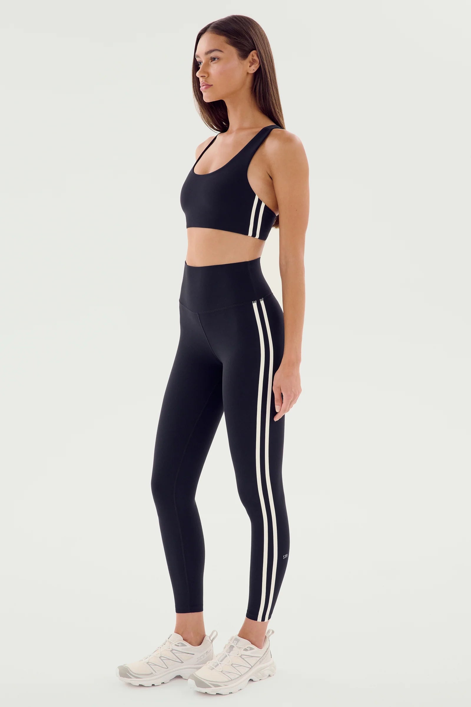 A woman stands in profile wearing a black sports bra and high-waisted leggings with white side stripes, and white athletic shoes, against a plain light background.