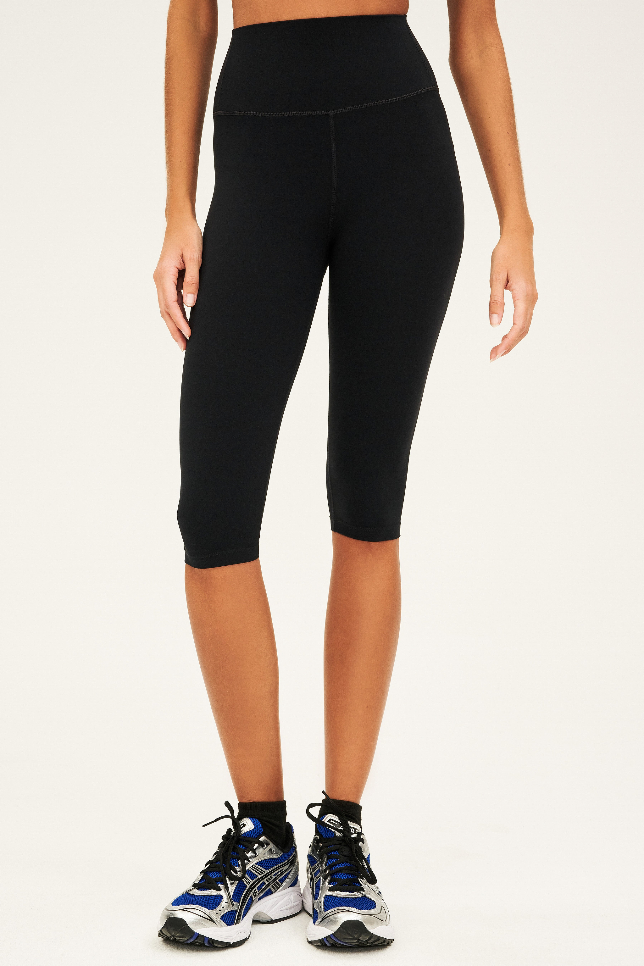 A person wearing black high-waisted, knee-length leggings and blue running shoes stands against a plain background.