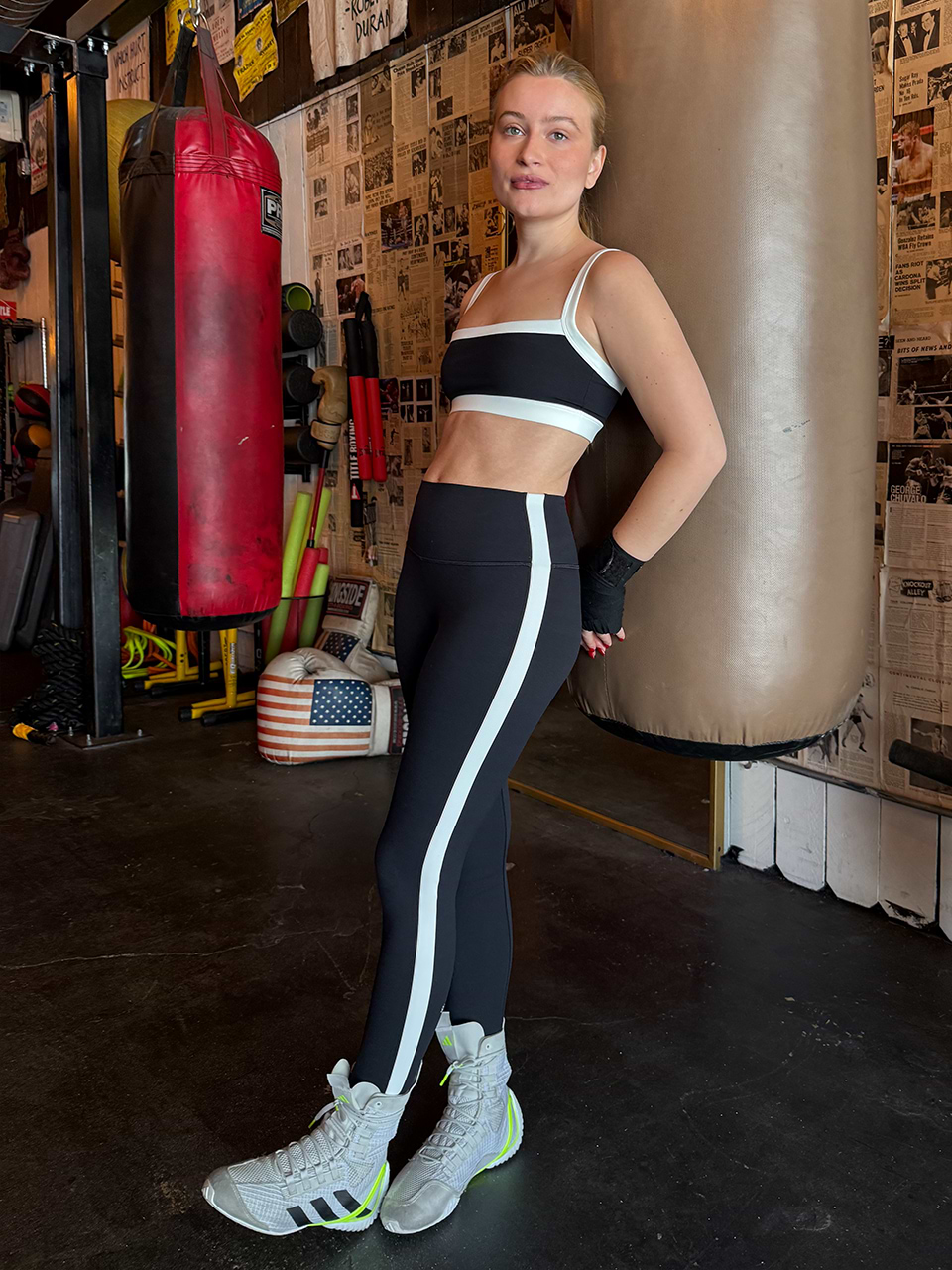 A woman in black and white athletic wear stands in a gym, leaning against a punching bag. Boxing gloves, a red bag, and gym equipment are visible in the background.