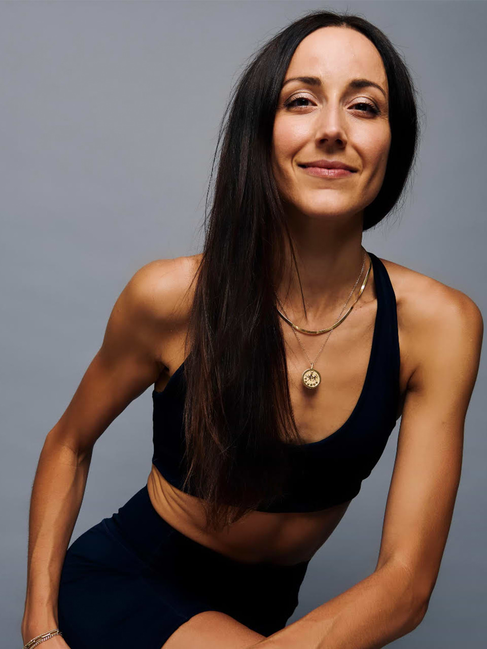 Woman with long dark hair wearing a black sports bra and necklace poses in front of a plain grey background, smiling slightly at the camera.