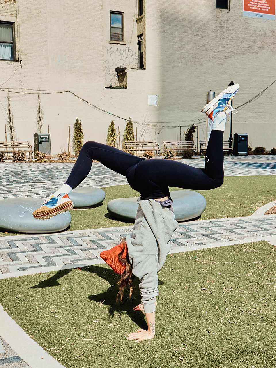 A person in athletic wear performs a handstand with bent legs on a grassy area in an urban outdoor setting.