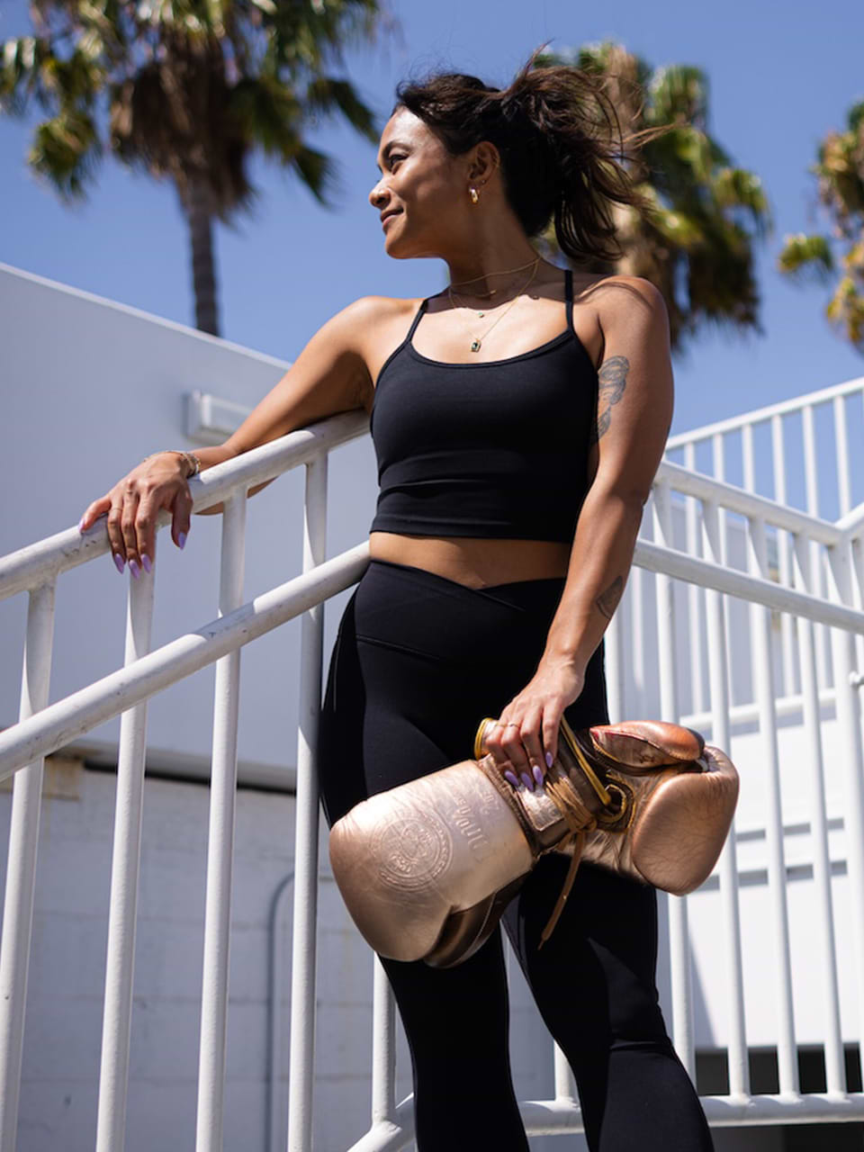 Woman in black athletic wear stands on an outdoor staircase, holding a pair of gold boxing gloves, with palm trees and blue sky in the background.