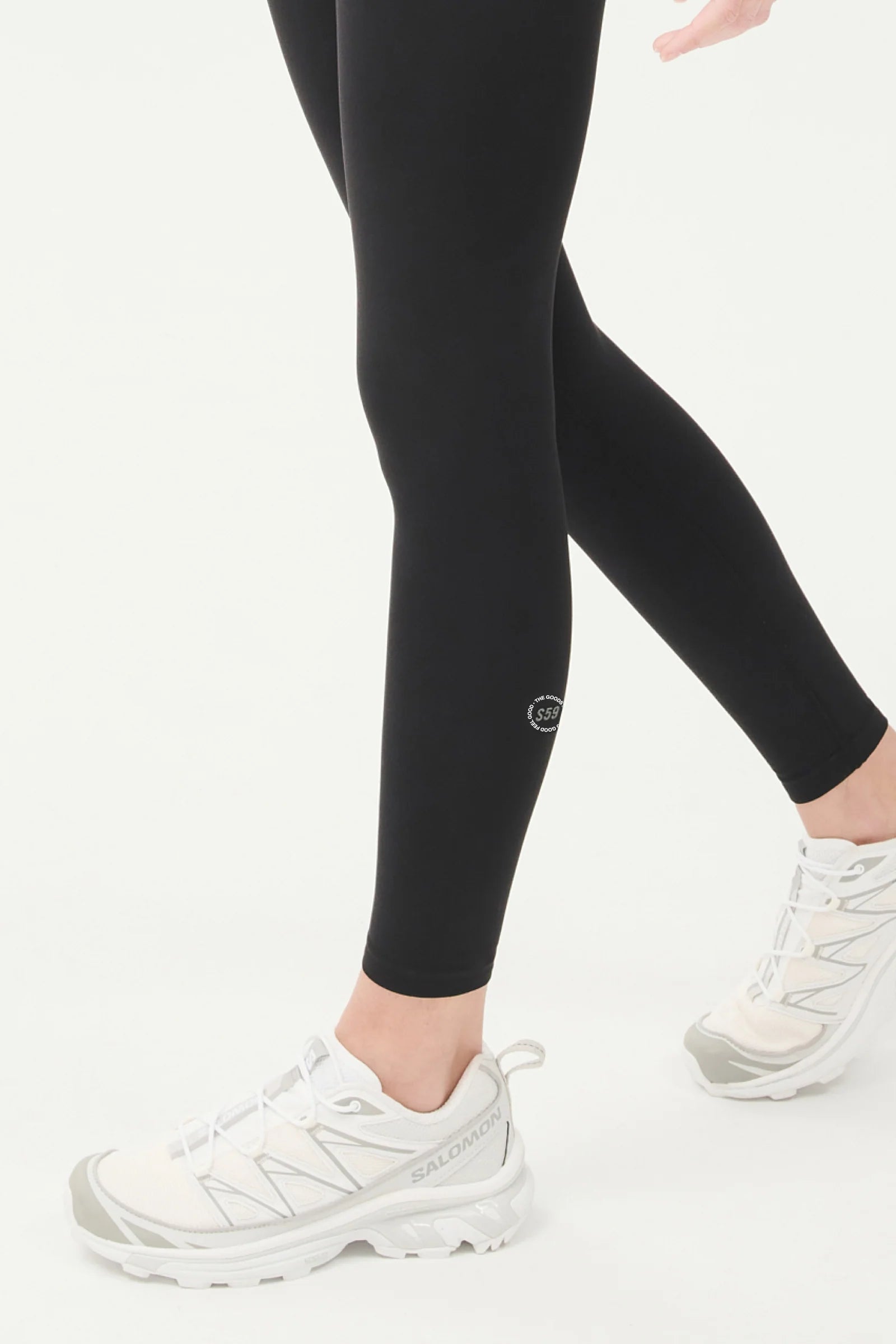 Person wearing black leggings and white Salomon athletic shoes, shown from the waist down, walking against a plain light background.