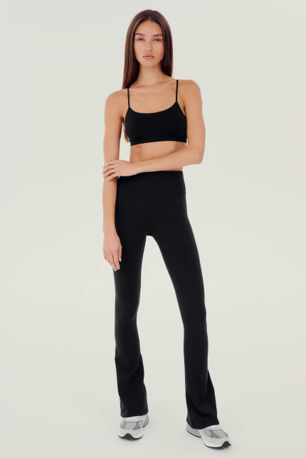 Woman standing against a plain background, wearing a black sports bra, black high-waisted flared leggings, and gray athletic shoes.