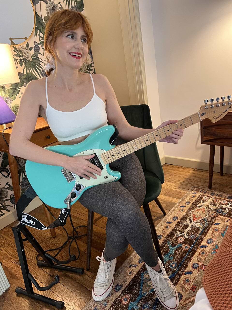 A woman sitting on a chair plays a teal electric guitar. She is wearing a white tank top, gray leggings, and white sneakers. The room has a patterned rug and wooden furniture.