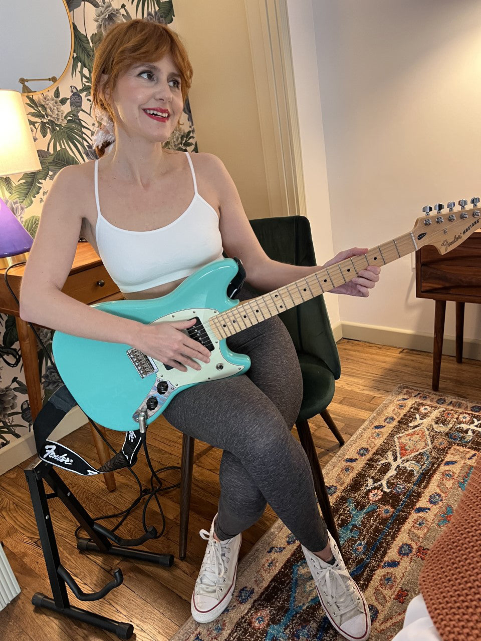 A woman sitting on a chair plays a teal electric guitar. She is wearing a white tank top, gray leggings, and white sneakers. The room has a patterned rug and wooden furniture.
