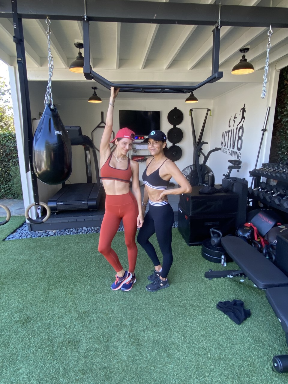 Two women in athletic wear stand and pose in a modern outdoor gym space with fitness equipment, weights, and a punching bag visible.