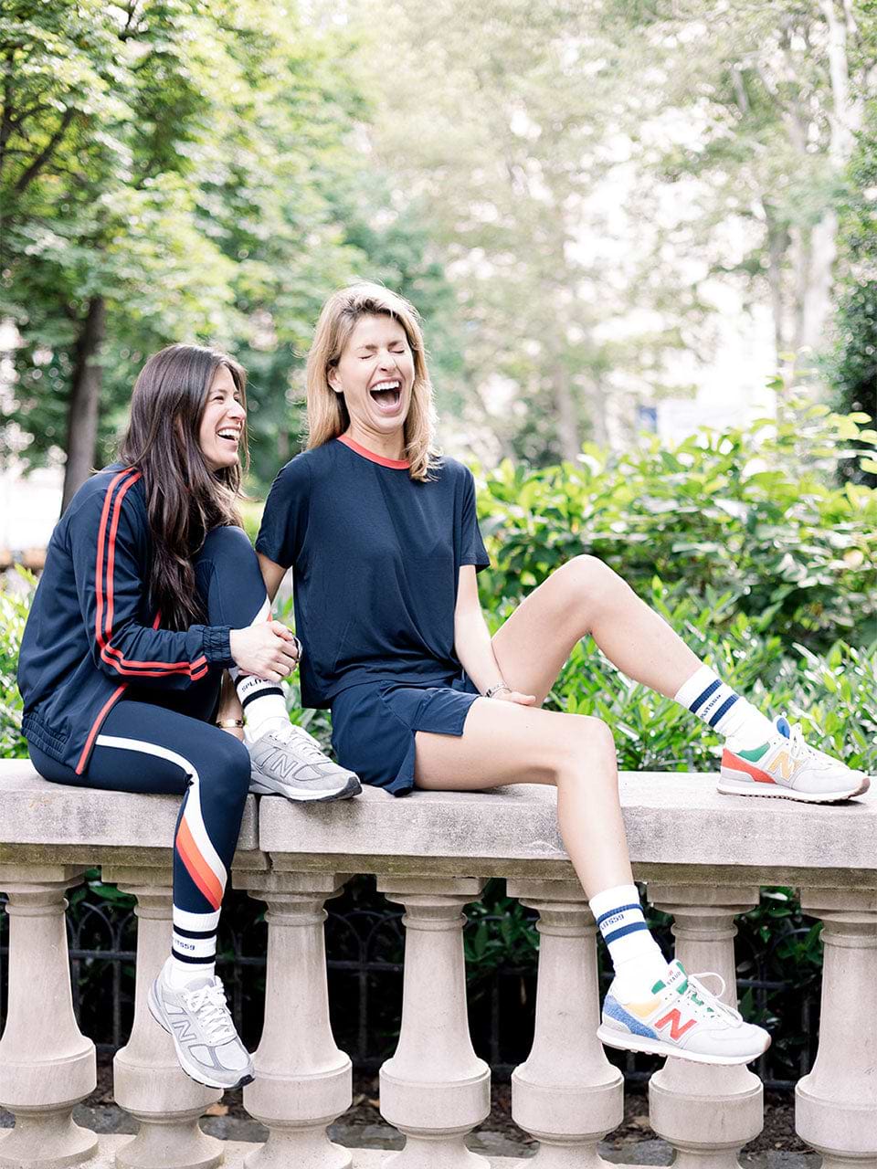 Two women in athletic clothing sit and laugh together on a stone bench outdoors, with greenery and trees in the background.