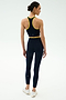 A woman faces away in the SPLITS59 Amber Airweight Bralette in Indigo/Ray, styled with matching leggings, featuring yellow trim and crafted from Airweight performance fabric, paired with white sneakers against a plain white background.