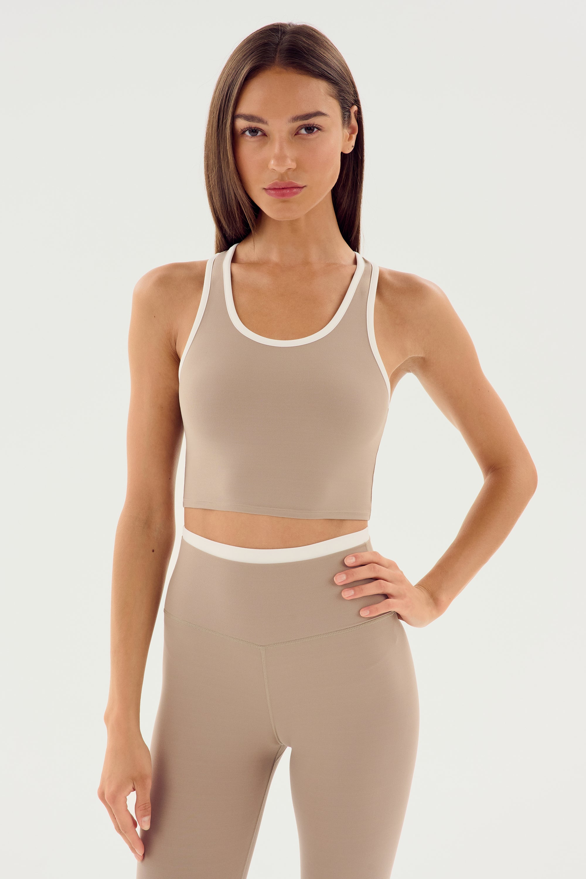A woman with long hair in a beige athletic outfit made from SPLITS59 Amber Airweight Bralette - Latte/White fabric stands with one hand on her hip against a plain white background.