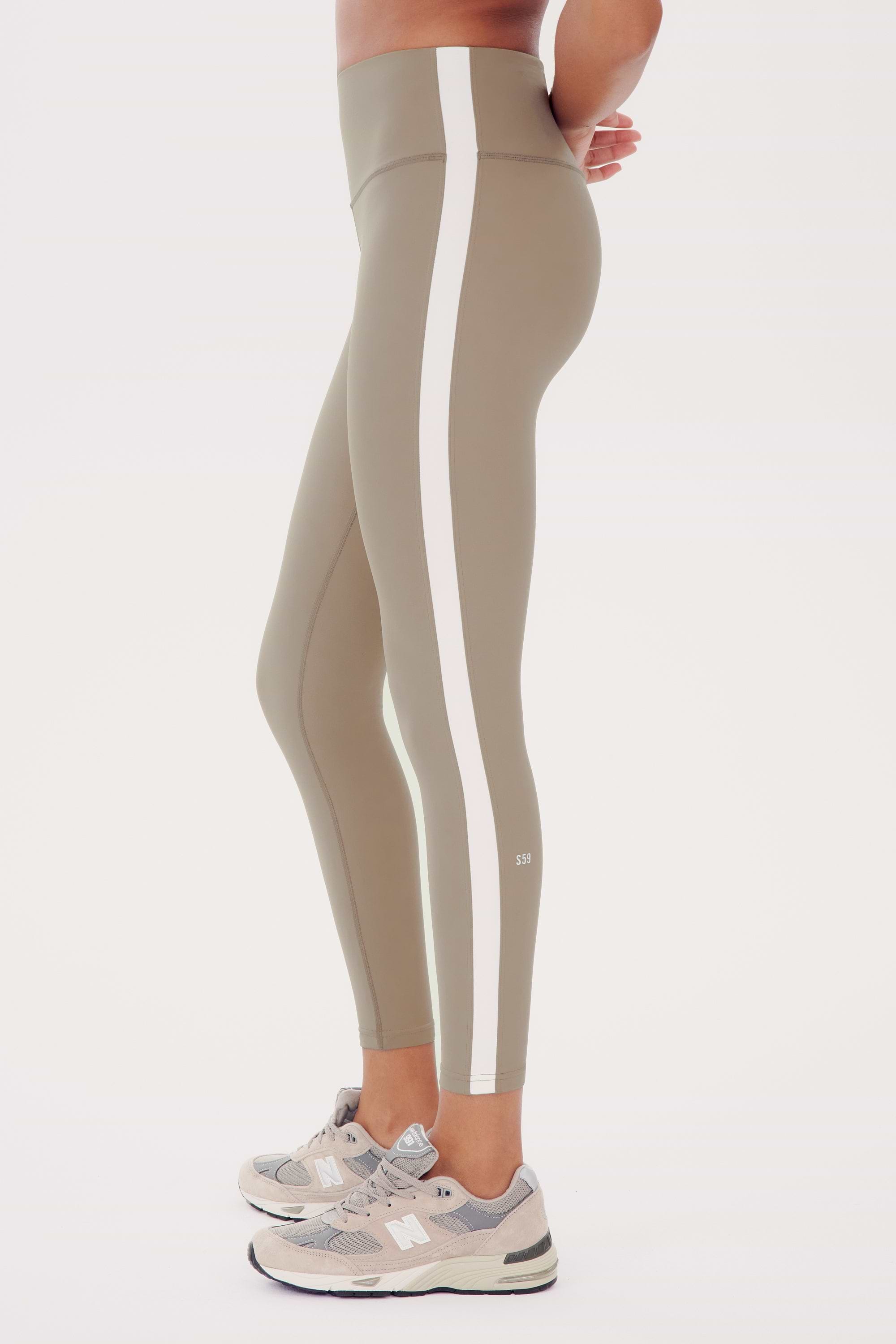 Side view of a woman in SPLITS59 Clare High Waist Rigor 7/8 - Latte/White high waist leggings with white side stripes, standing and showcasing the fit, paired with light grey sneakers.