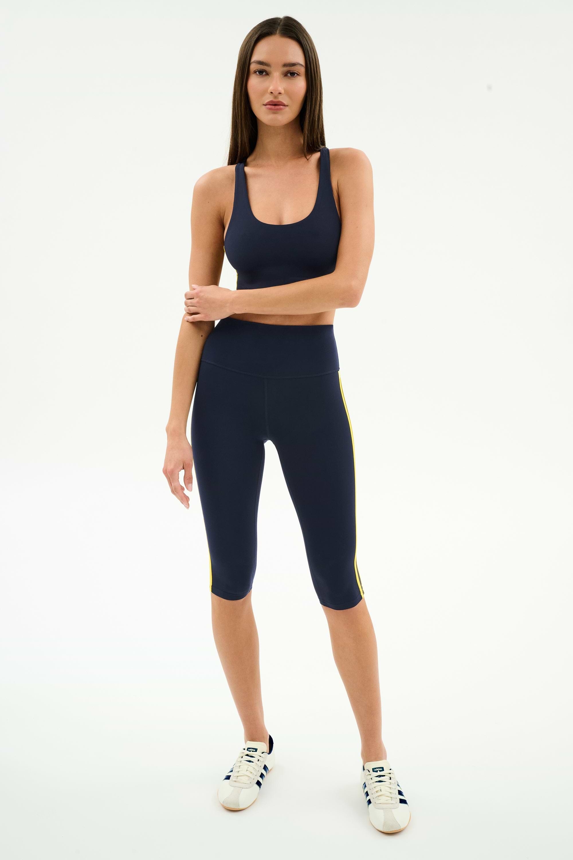 A woman stands facing forward in the SPLITS59 Ella Airweight Bra in Indigo/Bright Yellow, paired with matching capri leggings and white sneakers, all against a plain white background.