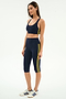 A woman wears the SPLITS59 Ella Airweight Bra in Indigo/Bright Yellow, featuring stretch nylon-spandex fabric and yellow side stripes, paired with white Adidas sneakers, posing against a plain white background.