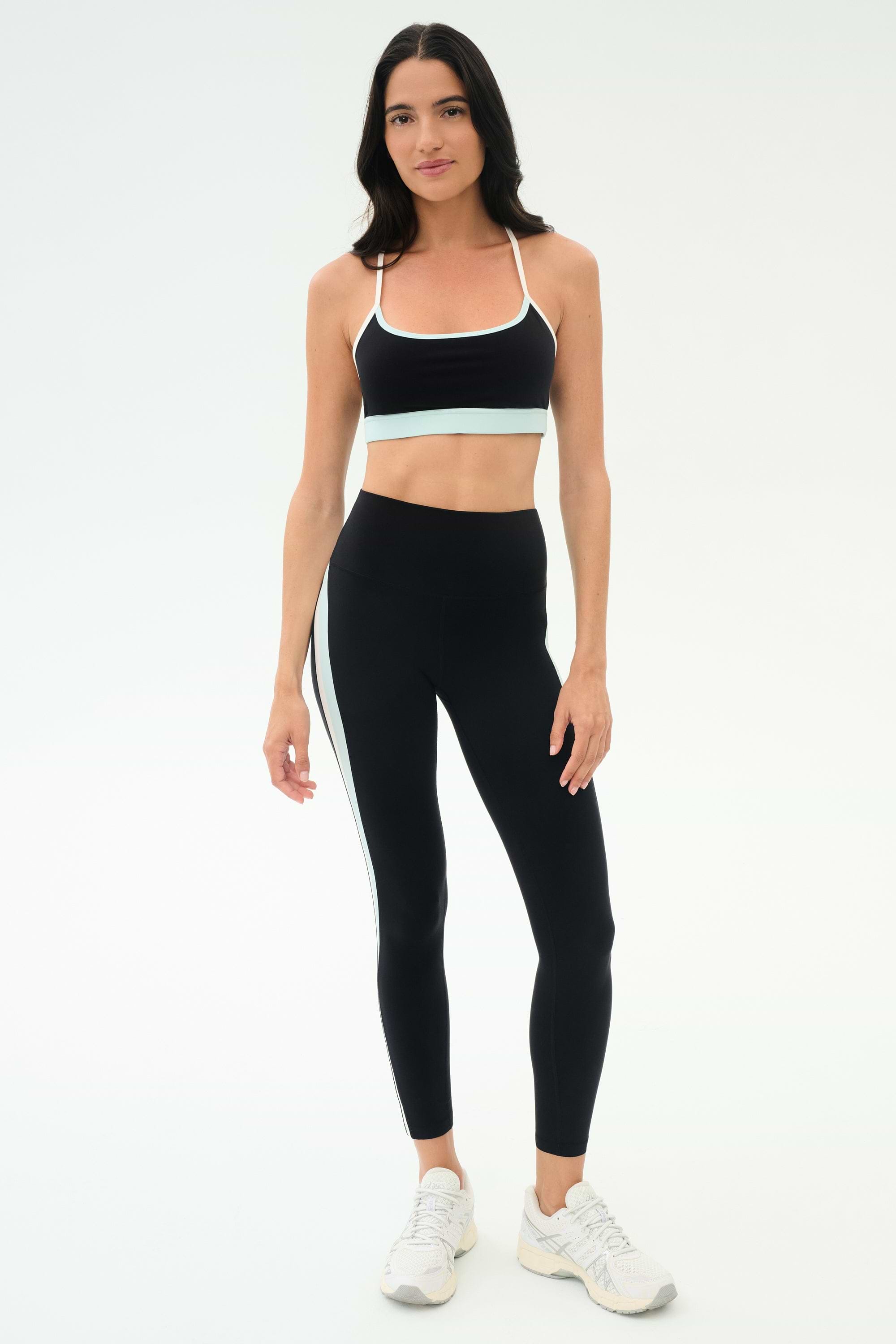 A woman stands against a plain background wearing the SPLITS59 Emerson Airweight Bra in Black/Ice, paired with matching black leggings featuring a white stripe and white athletic shoes—ideal for Pilates or hot yoga.