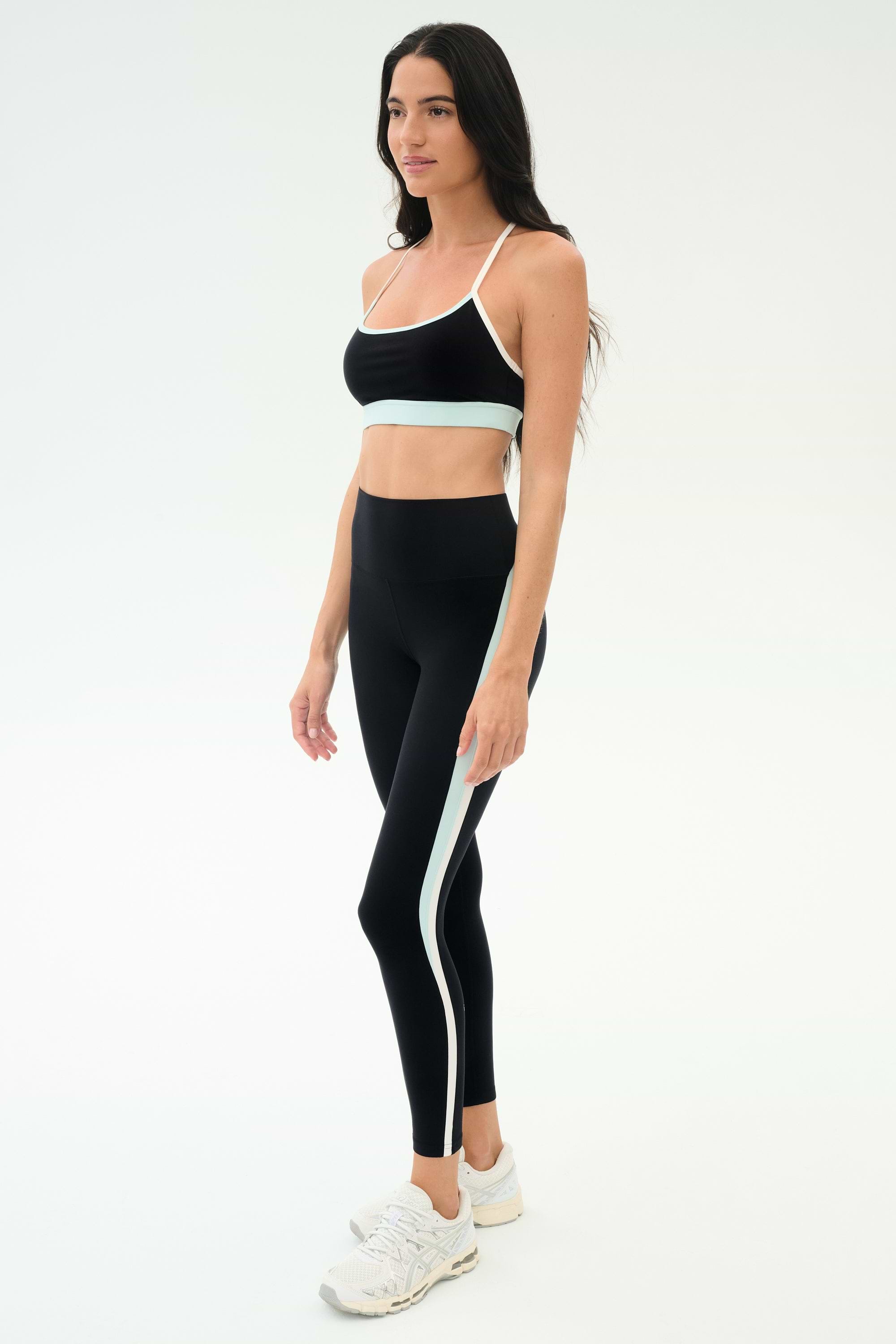 Woman in black SPLITS59 Emerson Airweight Bra - Black/Ice with white and light blue trim, standing slightly to the side against a plain white background—ideal for hot yoga or Pilates.