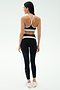 A woman faces away in a black SPLITS59 Emerson Airweight High Waist - Black/Ice legging with white accents and a black sports bra, paired with white sneakers—ideal for Pilates or hot yoga—against a plain white background.