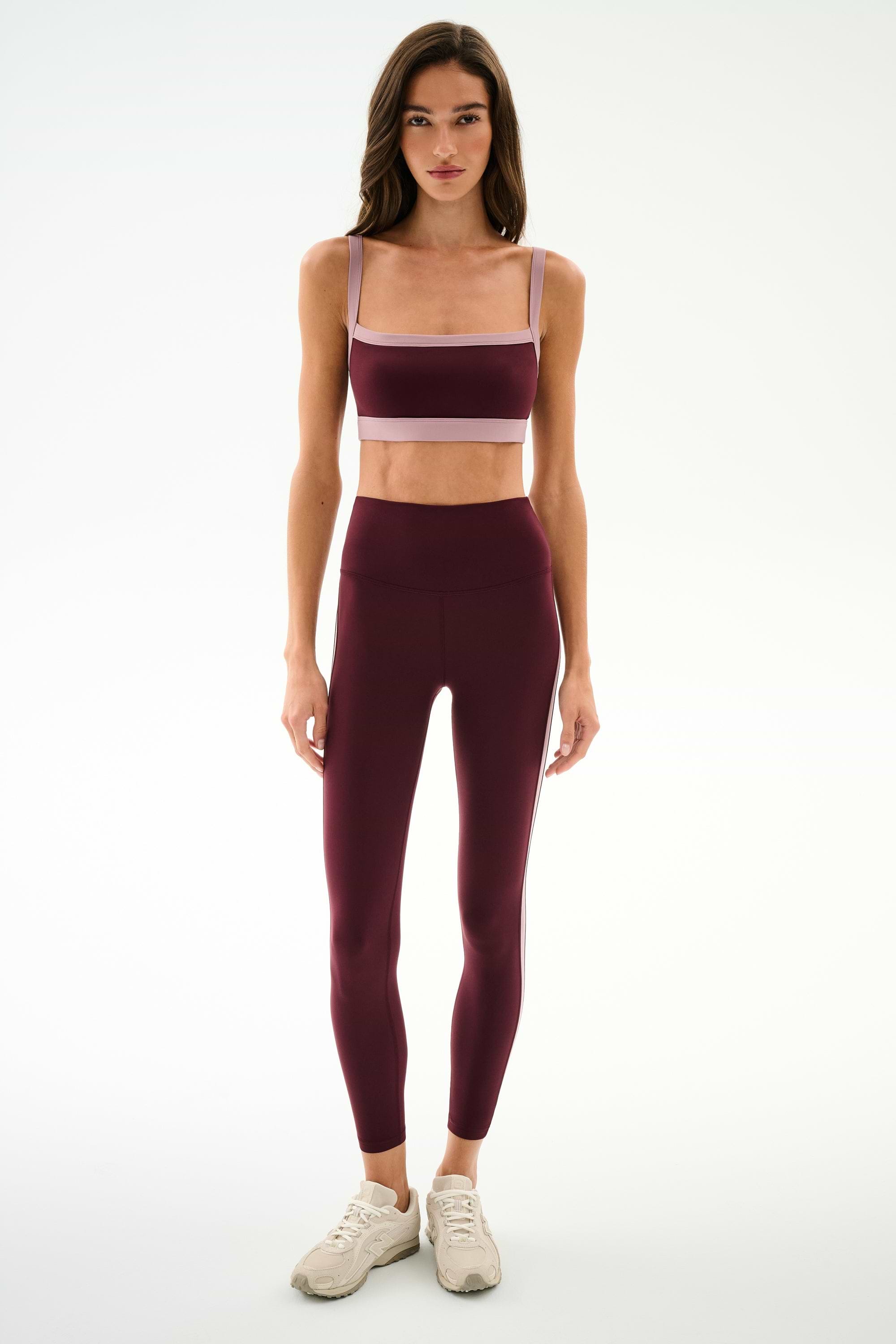 A person stands facing forward wearing the SPLITS59 Monah Rigor Bra in Dark Cherry/Blush with matching leggings and light-colored sneakers against a plain white background.